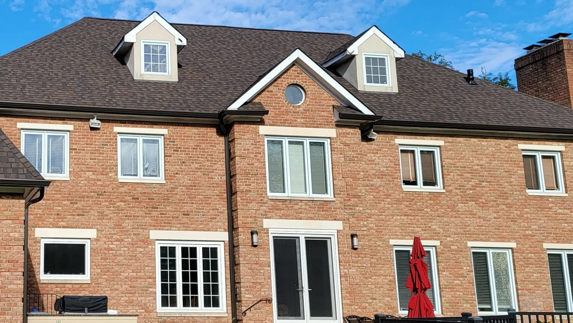 Large brick residential building with a dark asphalt shingle roof, dormer windows, white window trim, and a central gable, showing a well maintained traditional roofing installation.